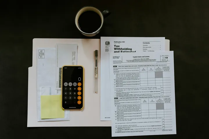 A workspace with tax forms, a calculator, a pen, an envelope, sticky notes, and a cup of coffee on a dark desk.