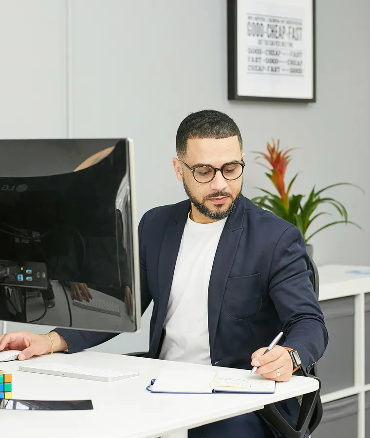 A man wearing glasses and a blazer working at a desk, writing in a notebook beside a computer monitor in a modern office.