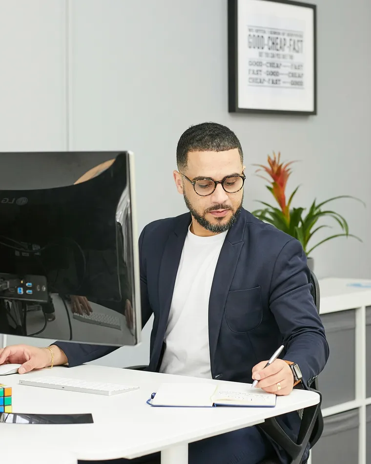 A man wearing glasses and a blazer working at a desk, writing in a notebook beside a computer monitor in a modern office.