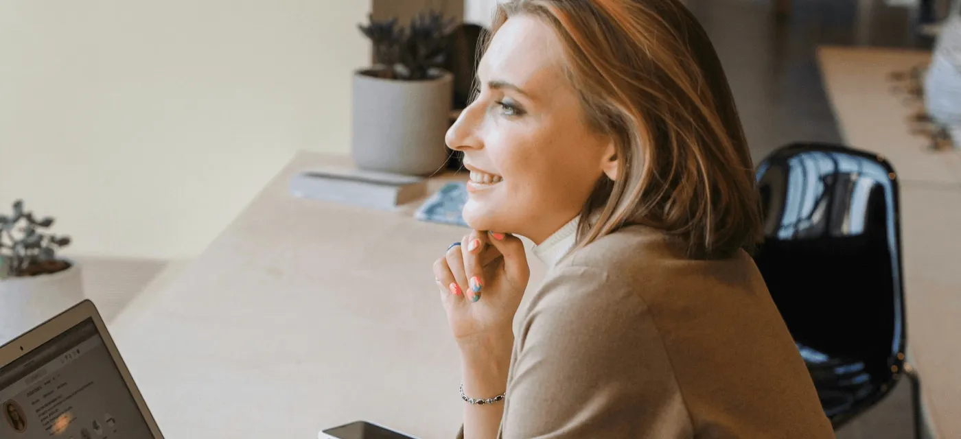 A woman in a brown coat smiling while working on a laptop in a bright workspace