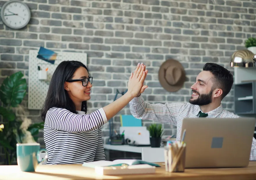 Two colleagues smiling and giving a high-five at a desk in a modern office with a laptop, coffee mug, and plants.